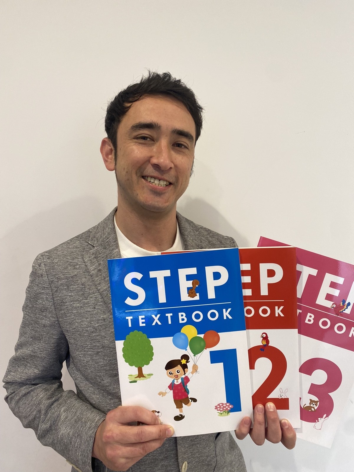 アルクの教材（STEP TEXTBOOK）を手に笑顔のネイティブ講師 Ty先生